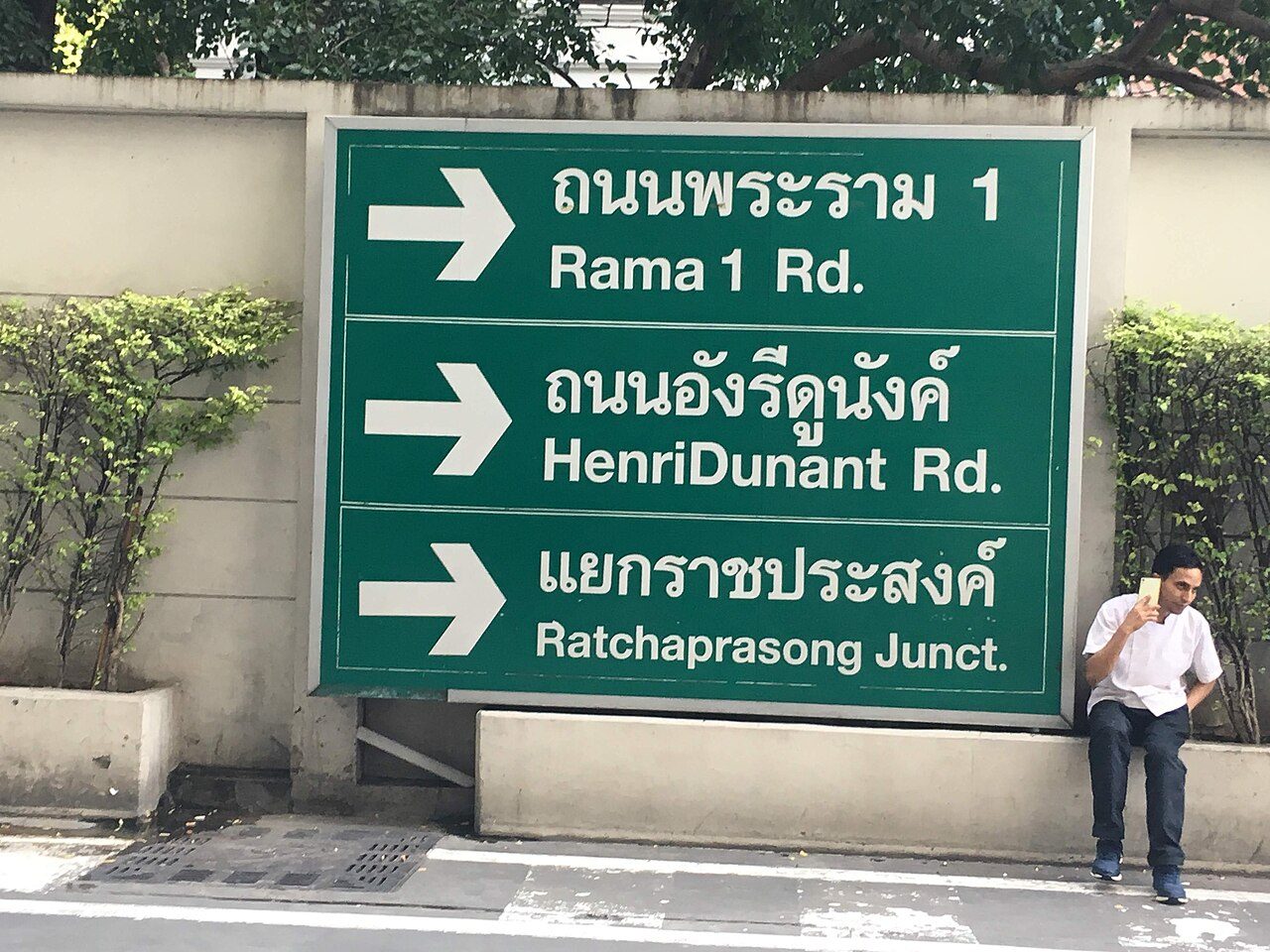 Green bilingual direction sign in Bangkok, Thailand, reading ถนนพระราม 1 (Rama 1 Rd.), ถนนอังรีดูนังต์ (Henri Dunant Rd.) and แยกราชประสงค์ (Ratchaprasong Junction) in Thai script above the English