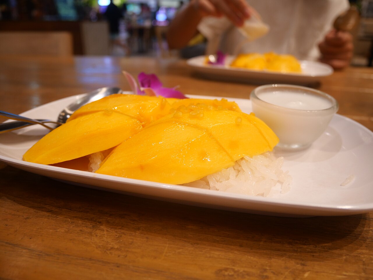 Plate of Thai mango sticky rice khao niao mamuang with sliced ripe mango, coconut sticky rice and a cup of coconut cream