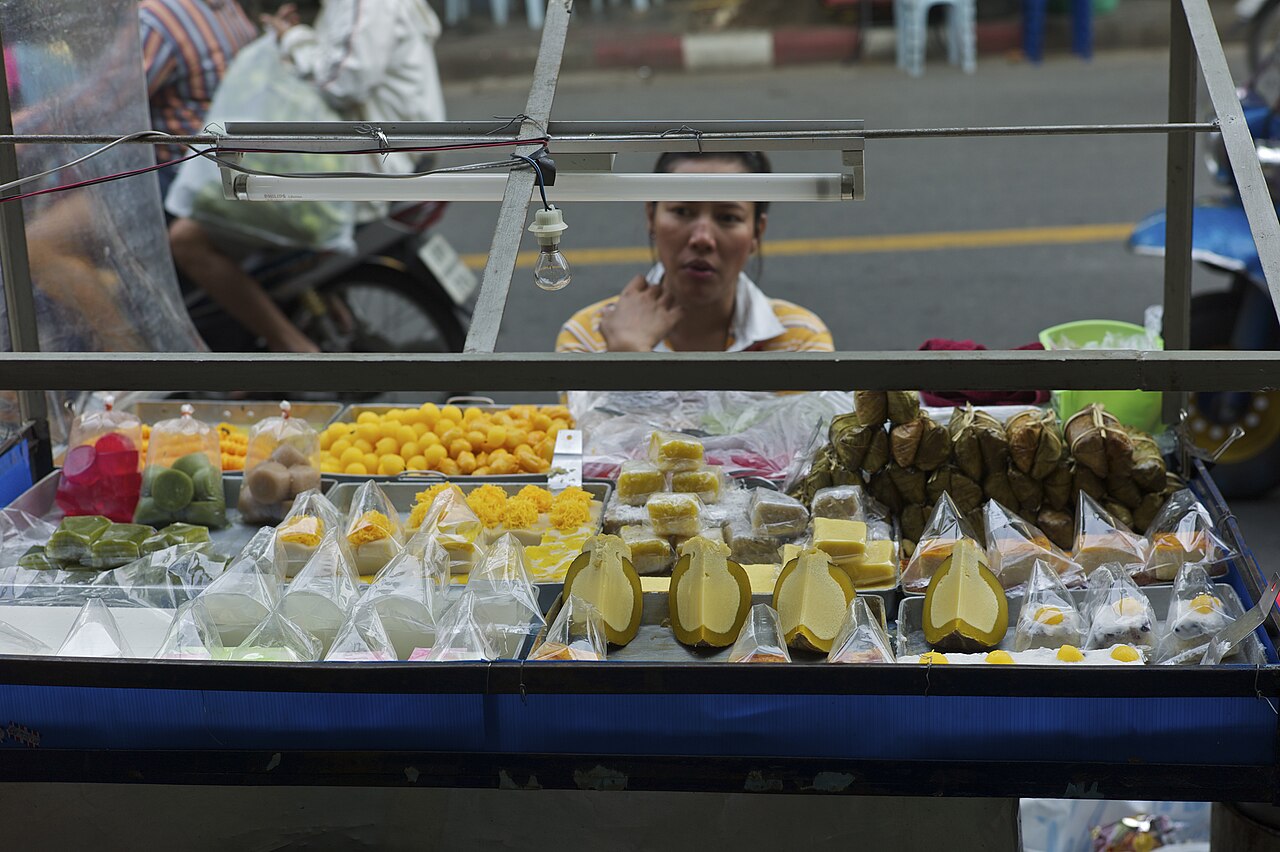 Bangkok street cart glass cabinet filled with Thai desserts including banana leaf parcels, yellow egg-yolk sweets and pandan-green squares