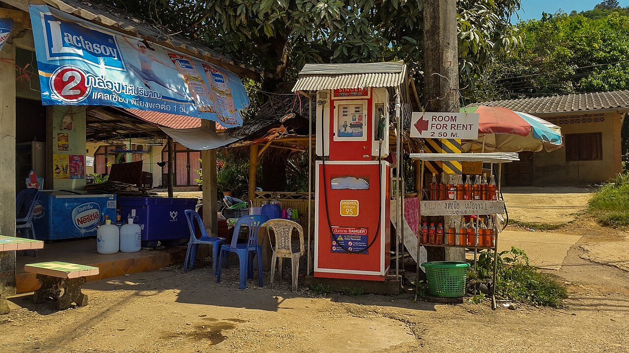 Single roadside gasohol pump on Koh Lanta, Thailand, with hand-lettered Thai price sign and glass bottles of petrol