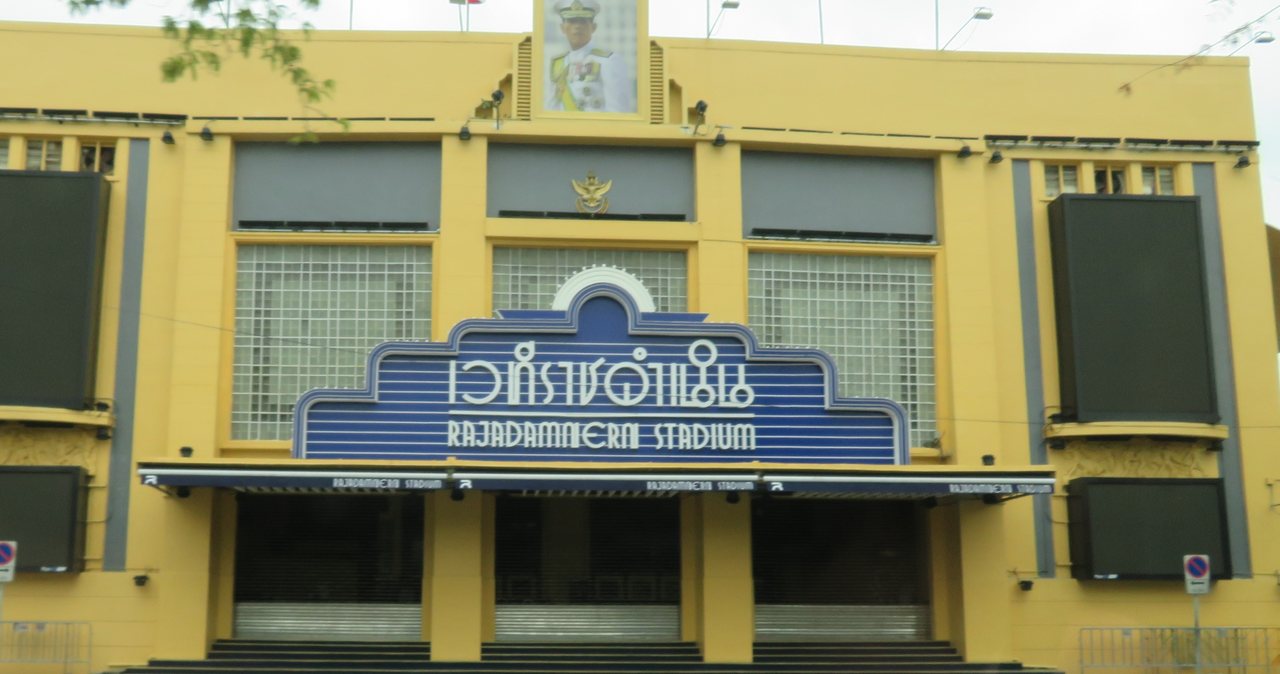 Thai script sign reading wethi Ratchadamnoen above the English words Rajadamnern Stadium on the yellow art deco facade in Bangkok