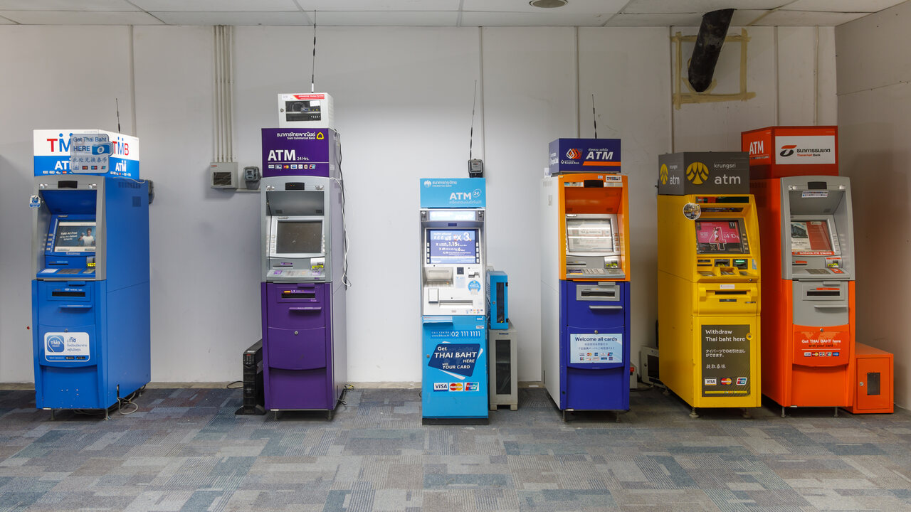 Row of six colour-coded Thai bank ATMs at Phuket International Airport in Thailand with bank names in Thai script above each machine