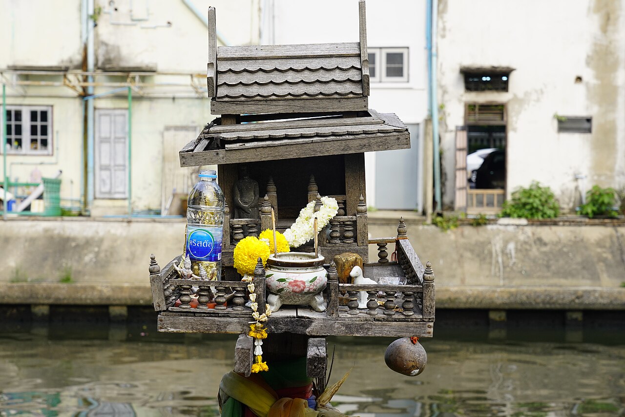 Weathered wooden Thai spirit house on a pillar above a canal in Phra Nakhon, Bangkok, Thailand, with garlands, incense pot and a water bottle offering