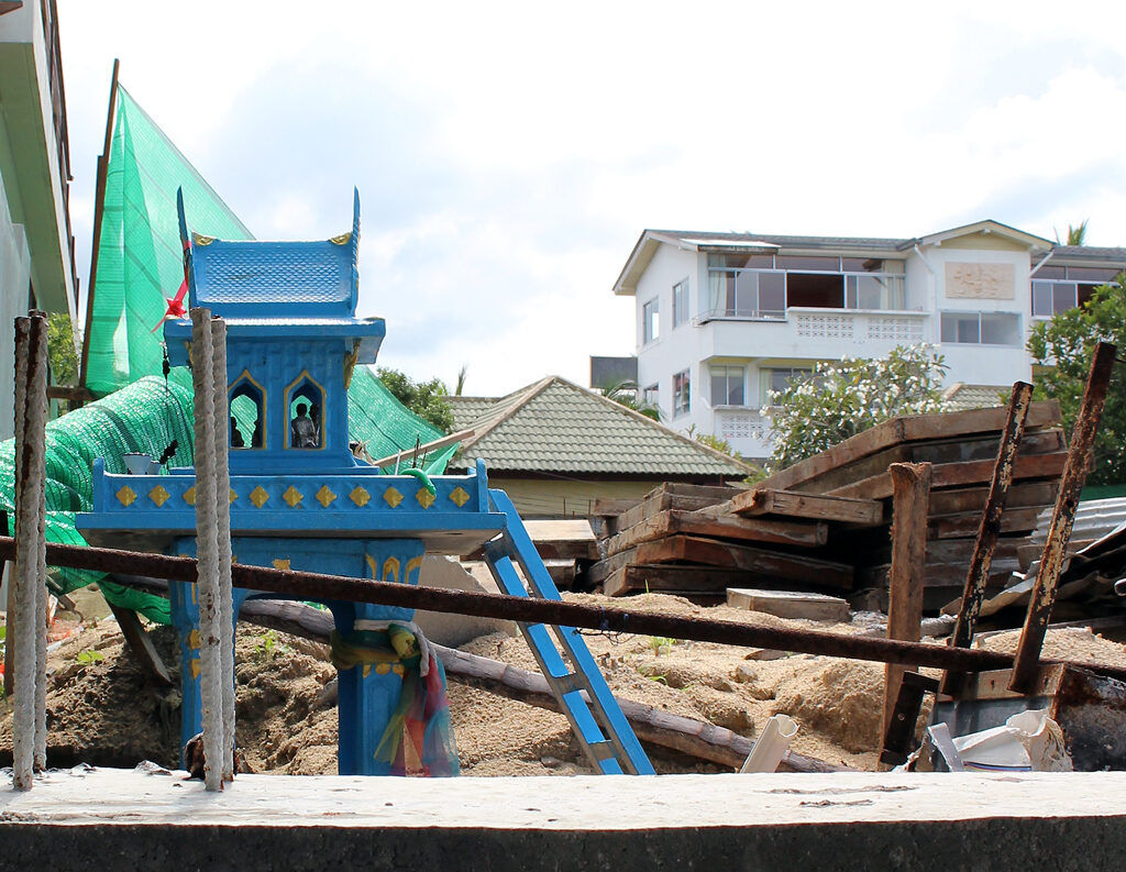 Blue Thai spirit house standing among rebar, timber and sand on a building site on Koh Samui, Thailand, with a coloured cloth tied around its pillar