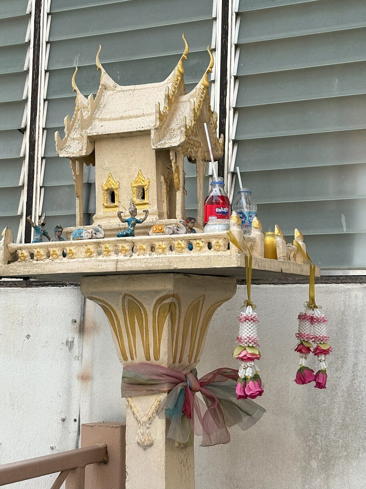 Close-up of offerings on a Thai spirit house in Chiang Rai, Thailand: a red Fanta bottle with a straw, bottled water, pink jasmine garlands and a coloured cloth tied round the pillar