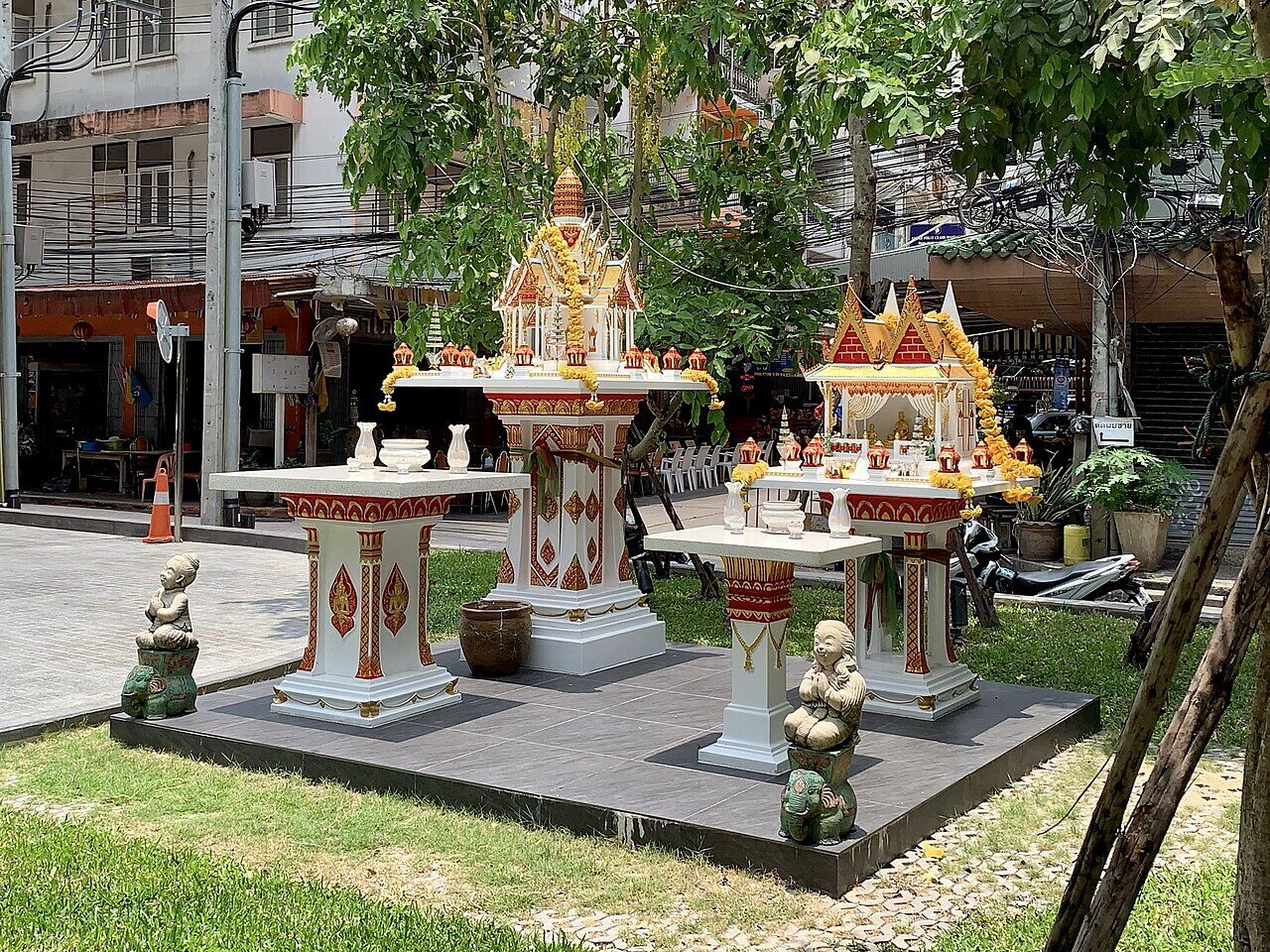 Two Thai spirit houses on a tiled platform outside a Bangkok hotel in Thailand, one tall single-pillar shrine and one lower house-shaped shrine with marigold garlands