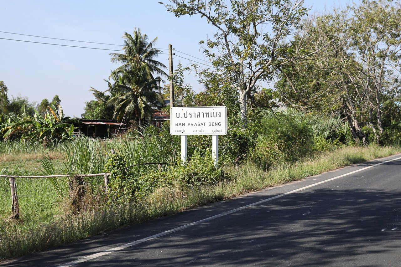 White village name sign beside a rural road in Surin Thailand reading Ban Prasat Beng in Thai script with English transliteration underneath