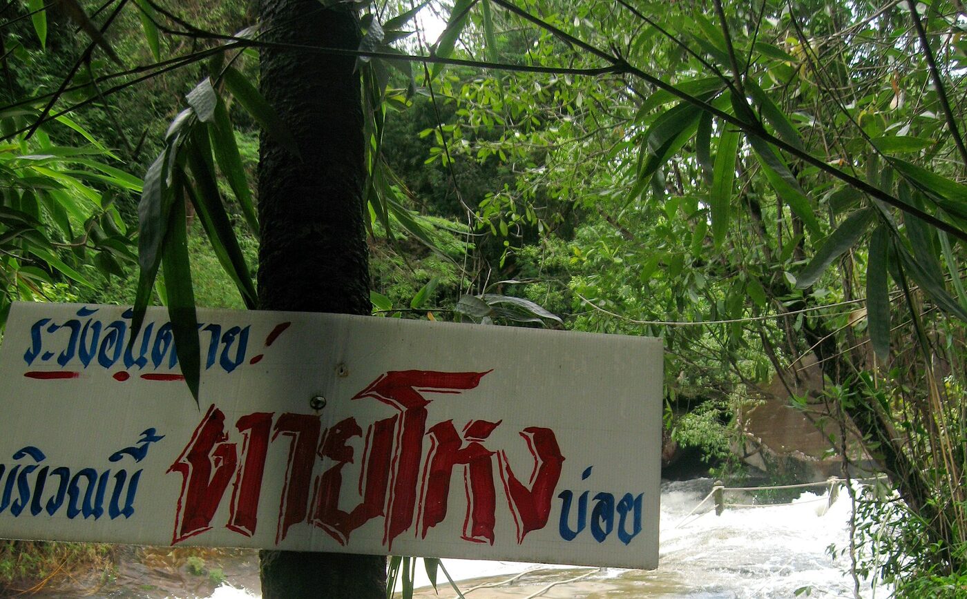 Hand-painted Thai warning sign in blue and red letters beside fast flowing water at a waterfall in northeastern Thailand