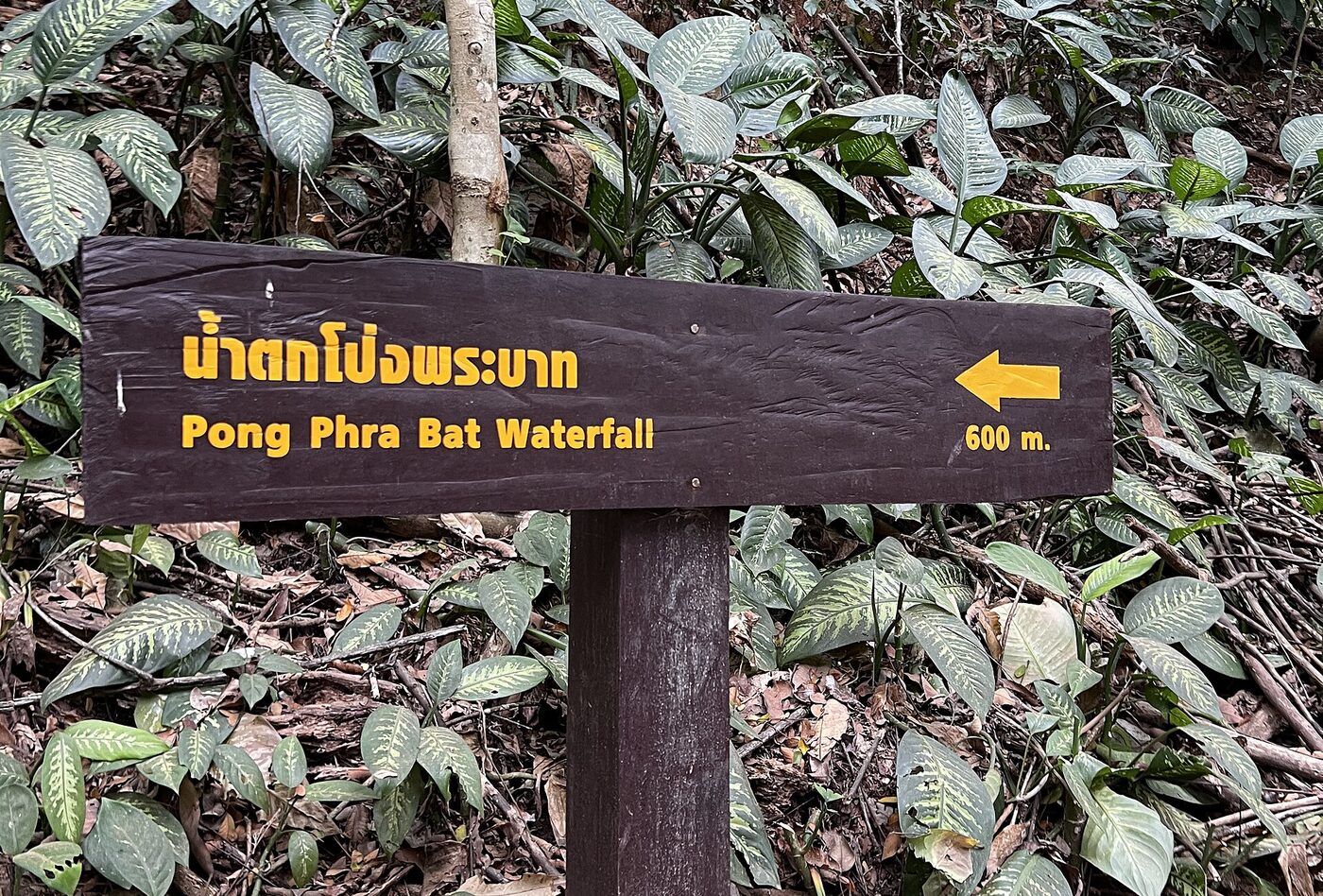 Brown wooden waterfall direction sign in Chiang Rai Thailand reading namtok Pong Phra Bat in yellow Thai letters with an arrow and 600 m