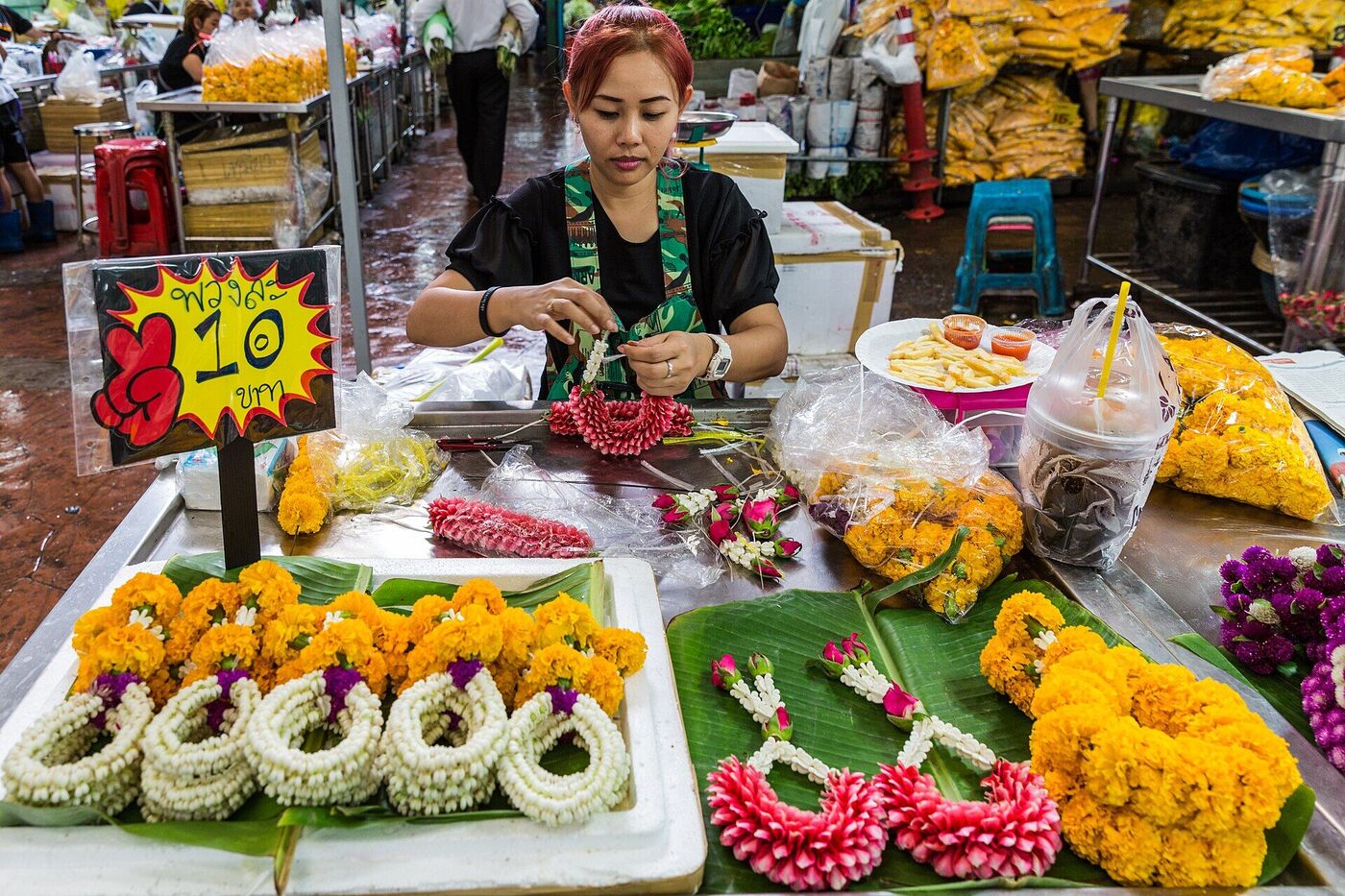 Thai vendor threading a jasmine flower garland at Pak Khlong Talat market in Bangkok Thailand beside a sign reading phuang la sip baht in Thai script
