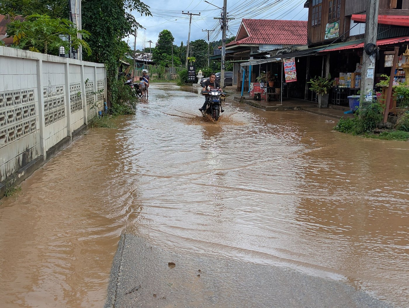 Motorcyclist riding through a flooded street past shophouses with Thai signs in Mae Sariang Thailand during the rainy season flood