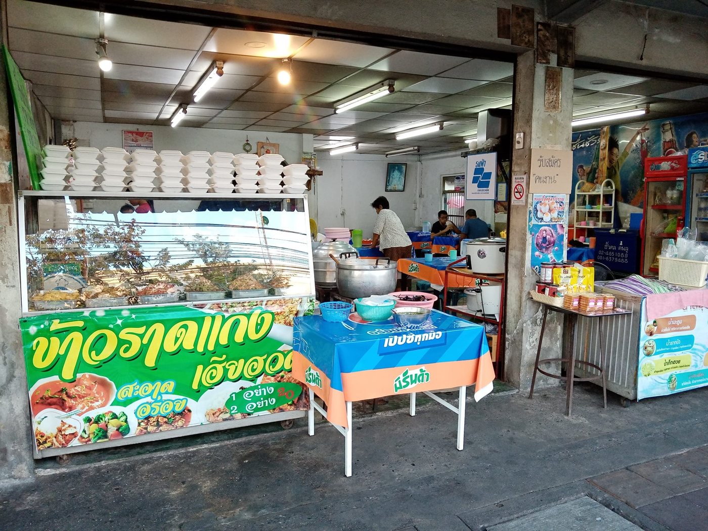 Thai rice and curry shop in Bangkok with a green banner reading khao raat kaeng in Thai letters, trays of curries in a glass cabinet and stacked takeaway boxes