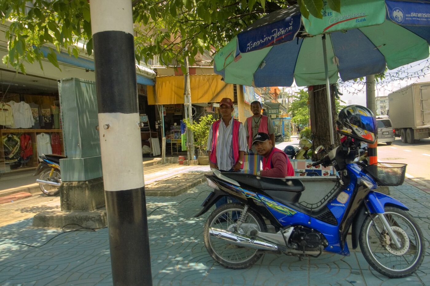 Thai motorbike taxi riders in pink vests waiting under a parasol beside a Honda Wave on a street in Cha-am, Thailand
