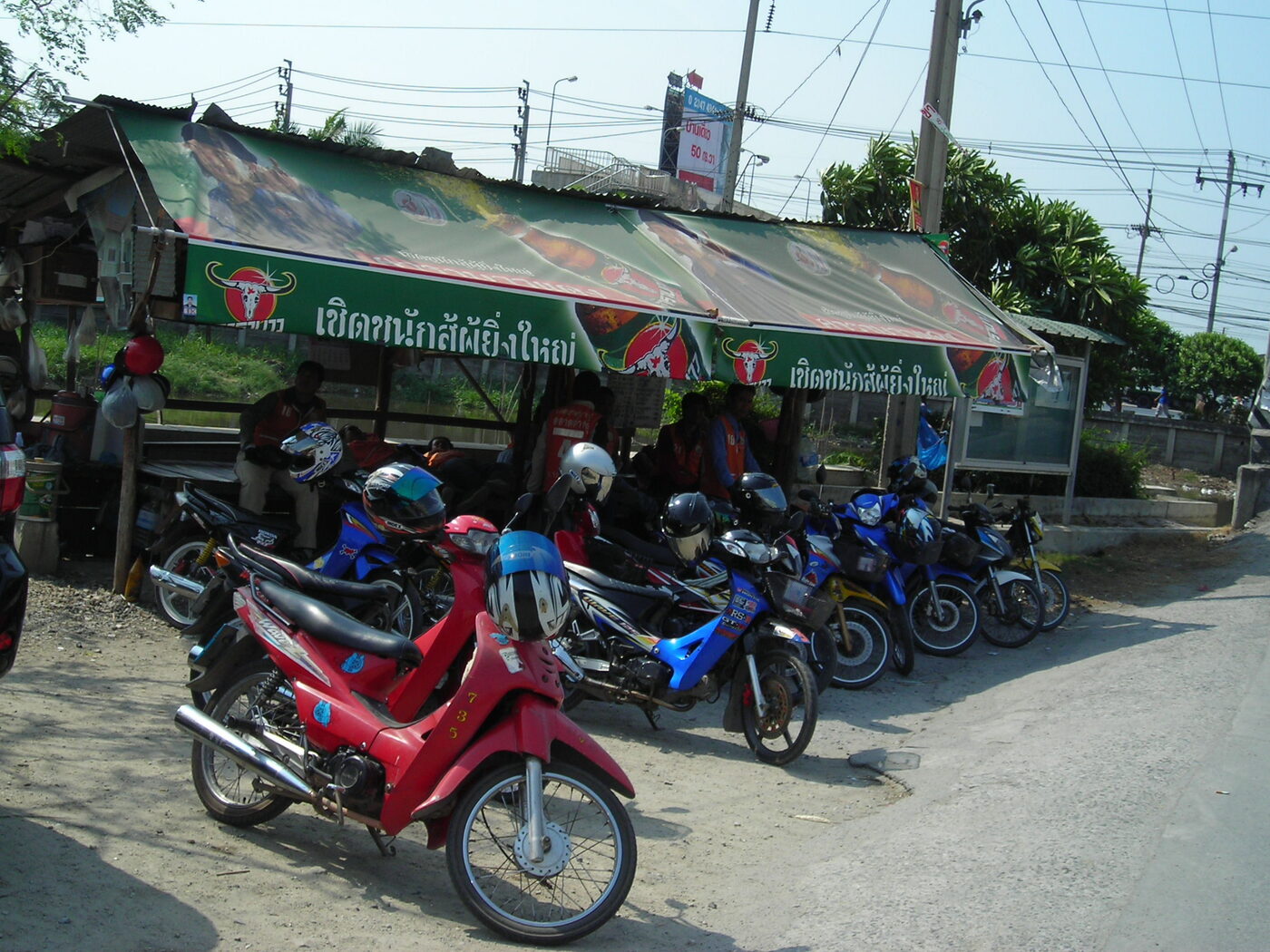 Thai motorbike taxi riders in orange vests waiting with their bikes and helmets under a roadside shelter in Samut Prakan, Thailand