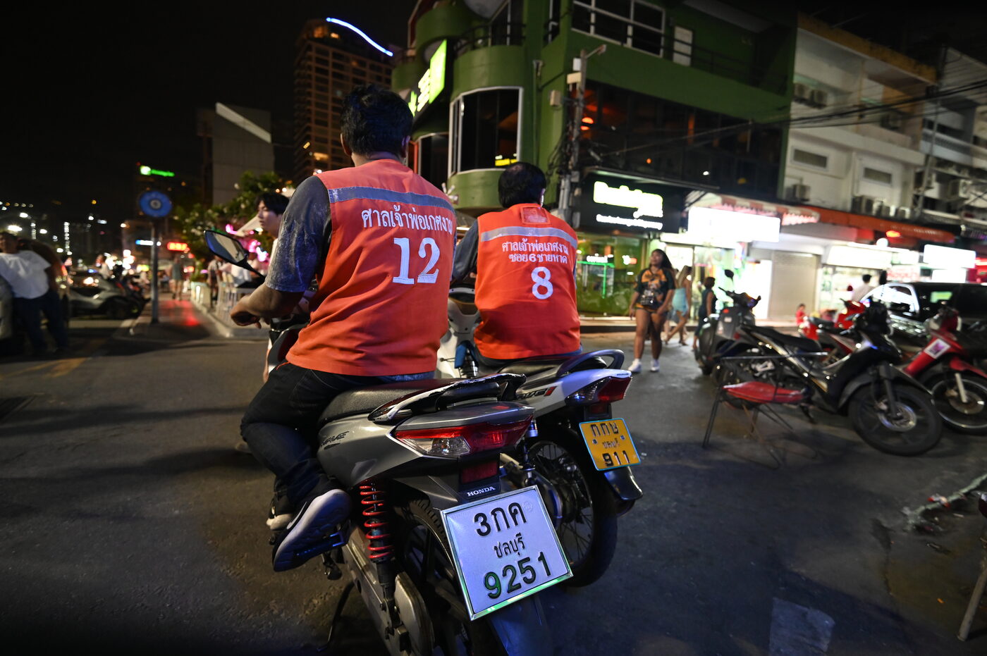 Two Thai motorbike taxi riders in numbered orange vests with Thai script, waiting at a street corner in Pattaya, Thailand