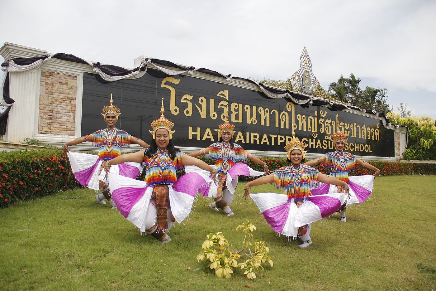 Students in Thai classical dance costume in front of a Thai school sign in Hat Yai with gold Thai script above its English transliteration