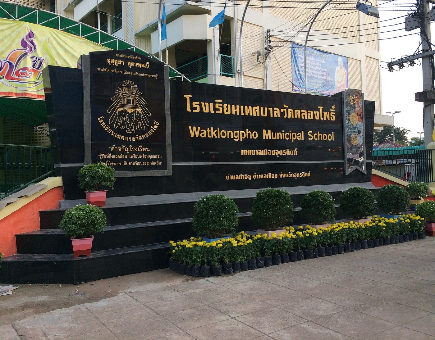 Thai school sign in gold letters on black granite at a municipal school gate in Uttaradit, Thailand, with Thai script, English romanisation and the district address line