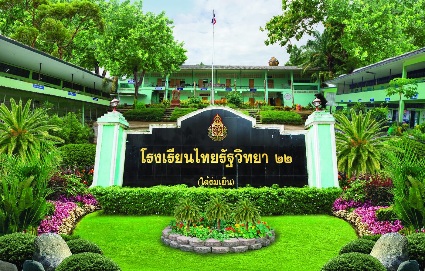 Black and gold Thai school sign reading Thairath Witthaya 22 with a Thai numeral, in front of green school buildings in Thailand