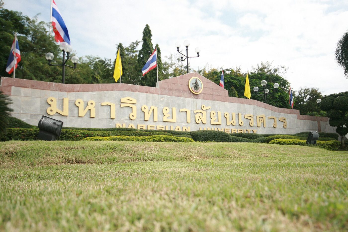 Large gold Thai letters reading Naresuan University on a stone wall at the campus entrance in Phitsanulok, Thailand, with Thai flags behind