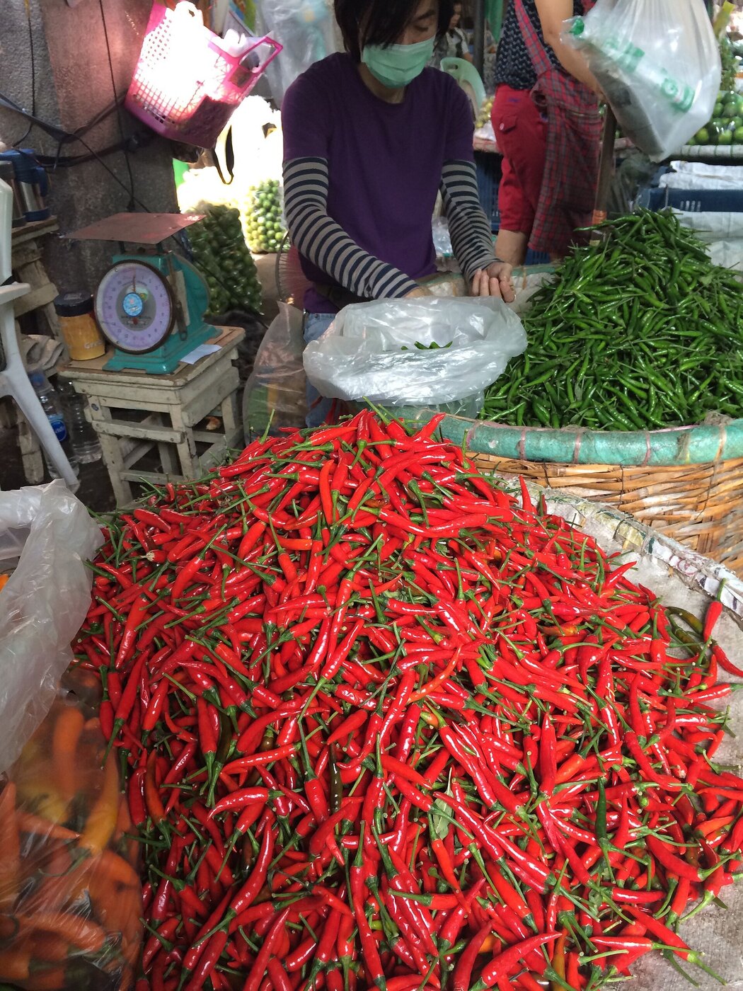 Vendor behind a huge pile of fresh red and green chillies with weighing scales at a market stall in Bangkok Thailand, illustrating Thai vocabulary for prik chillies