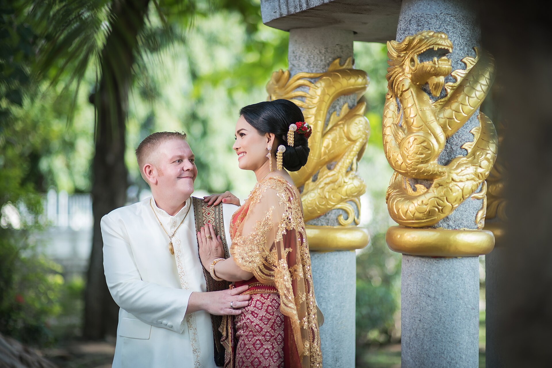 Thai bride in gold chut thai and foreign groom posing for pre-wedding photographs beside naga pillars at a temple in Thailand