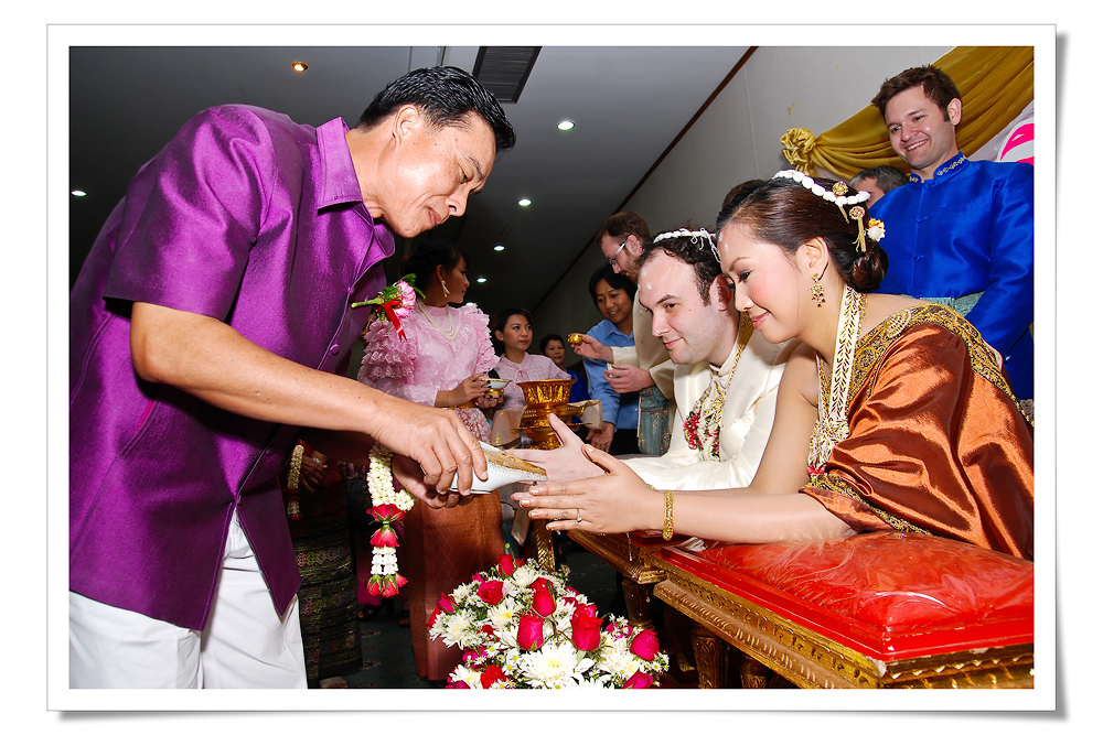 Elder pouring conch shell water over the hands of a bride and groom wearing mongkhon headbands at a rod nam sang Thai wedding ceremony