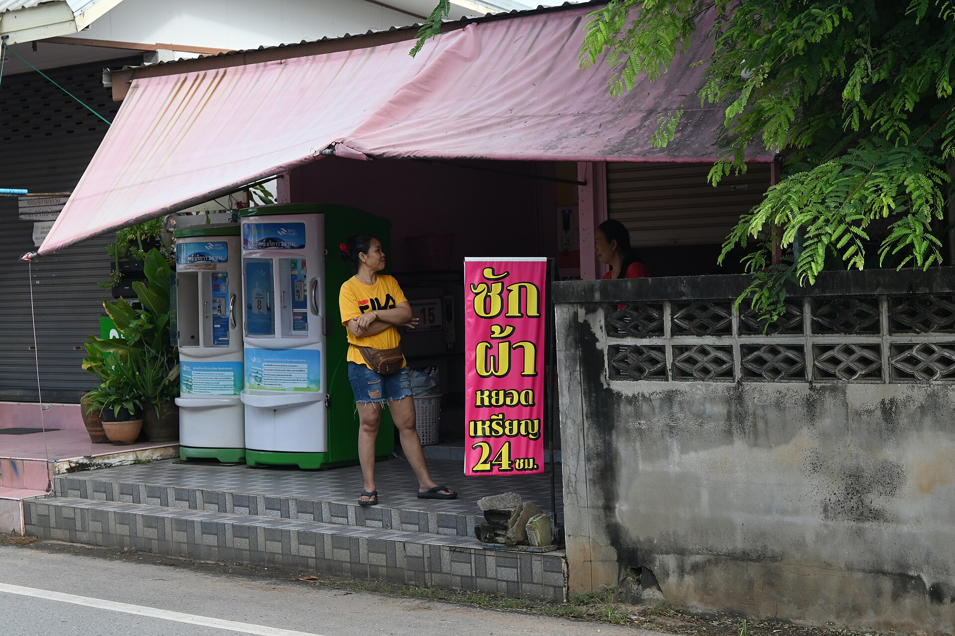 Roadside Thai laundry shop with coin-operated washing machines and a pink sign reading sak pha yot rian 24 chom in Thai script