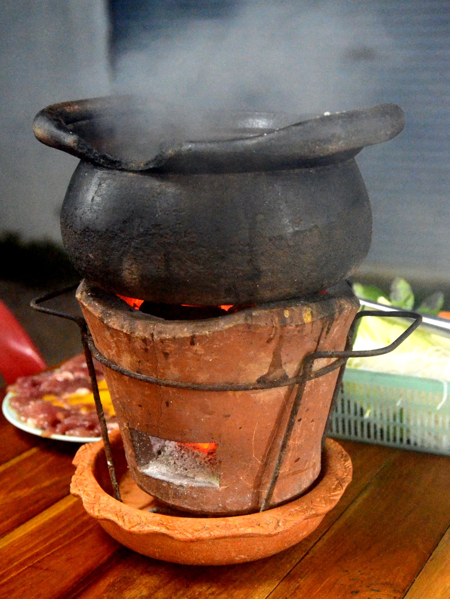 Blackened clay pot of chim chum herbal hot pot steaming on a terracotta charcoal brazier beside a plate of marinated meat in Thailand