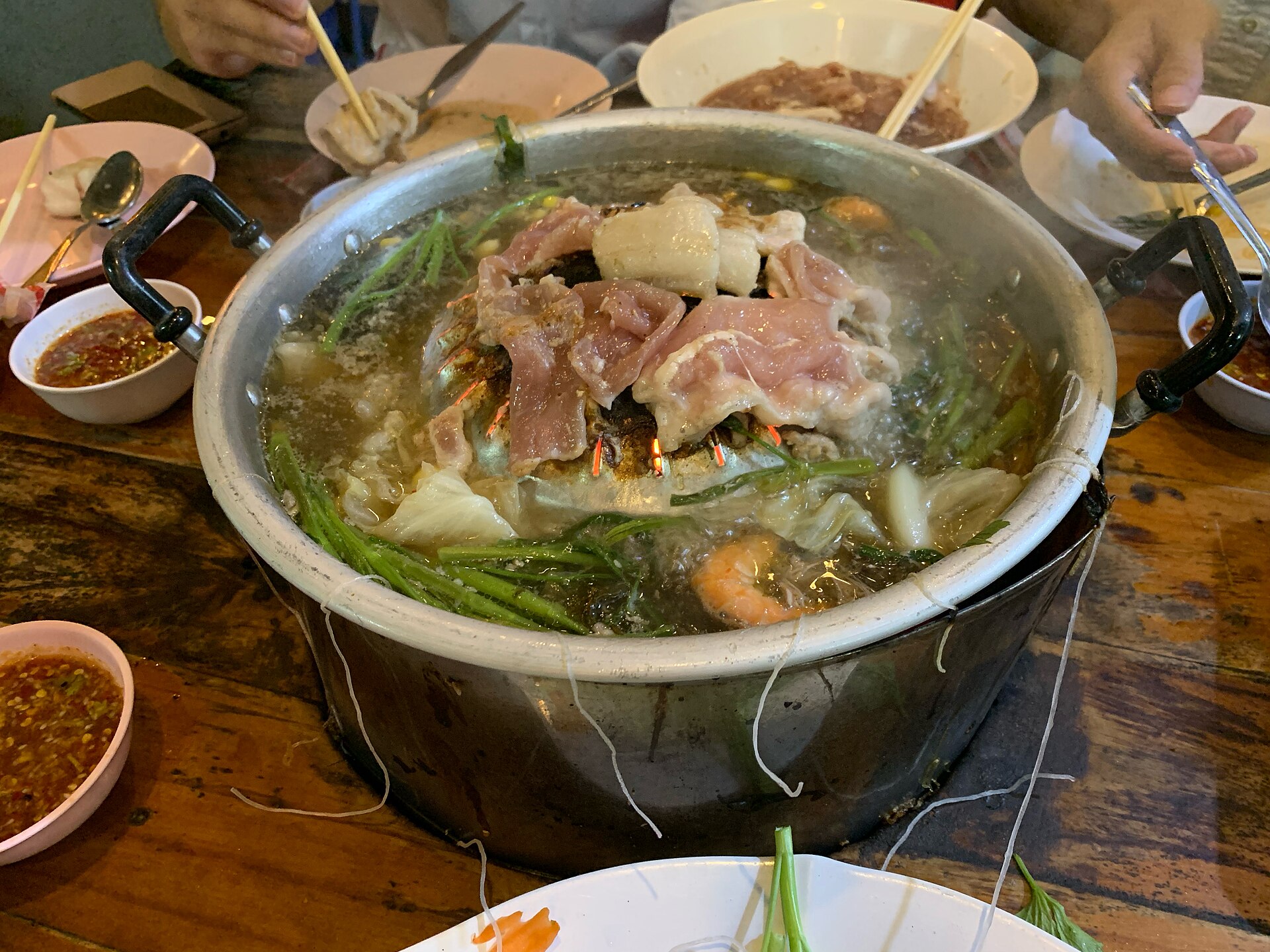 Mookata pan with a domed grill centre, raw pork slices and a broth moat of vegetables and glass noodles on a wooden table in Nakhon Nayok, Thailand