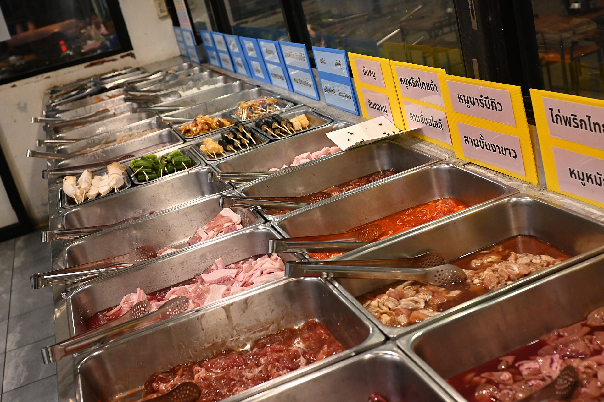 Thai BBQ buffet counter in Thailand with stainless steel trays of marinated pork and chicken under yellow and blue cards labelled in Thai script