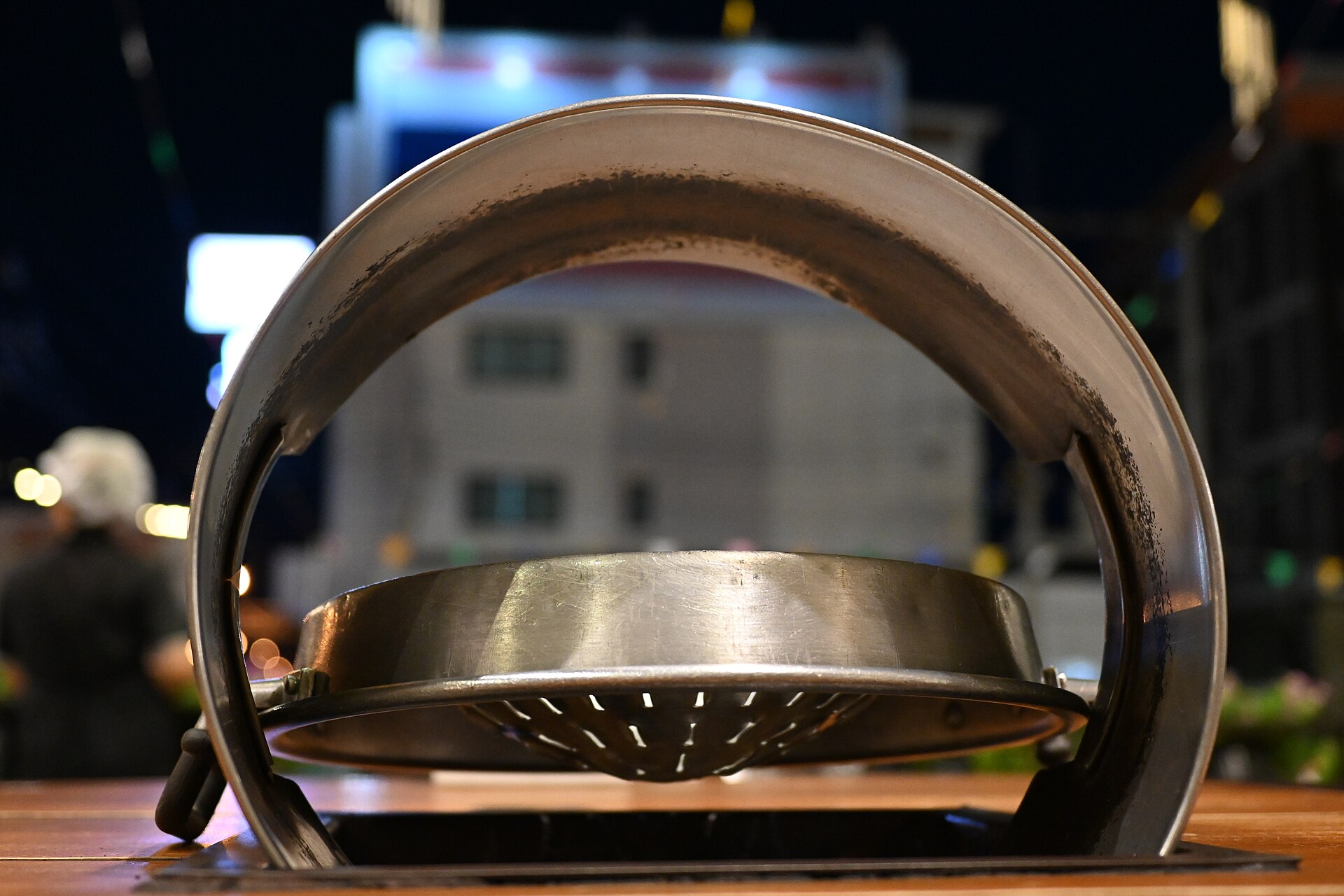 Empty domed Thai BBQ grill pan seen edge-on above a charcoal burner set into a wooden table at a street-side mookata restaurant in Thailand at night