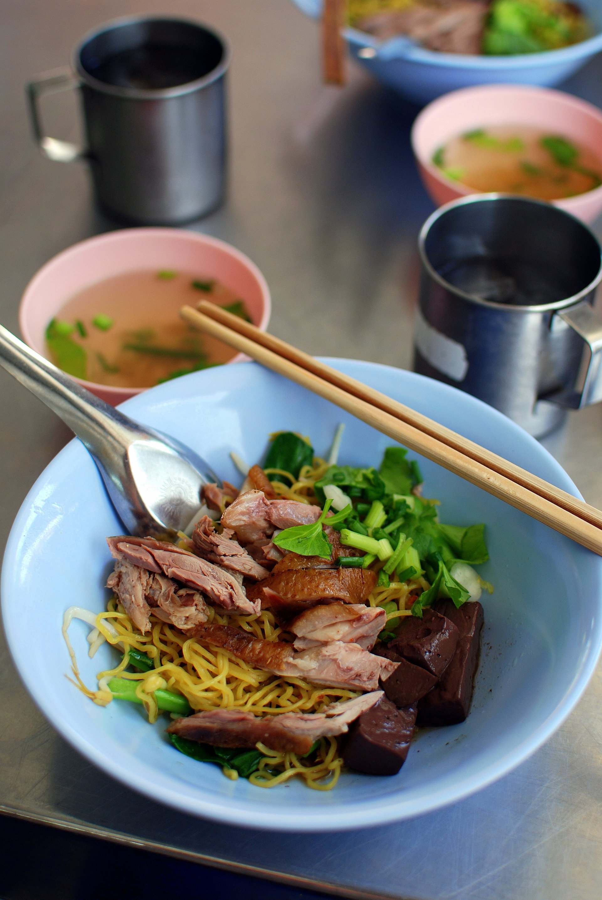 Bowl of Thai bamee haeng dry egg noodles topped with sliced duck, blood cubes and greens, with a separate pink bowl of clear broth beside it