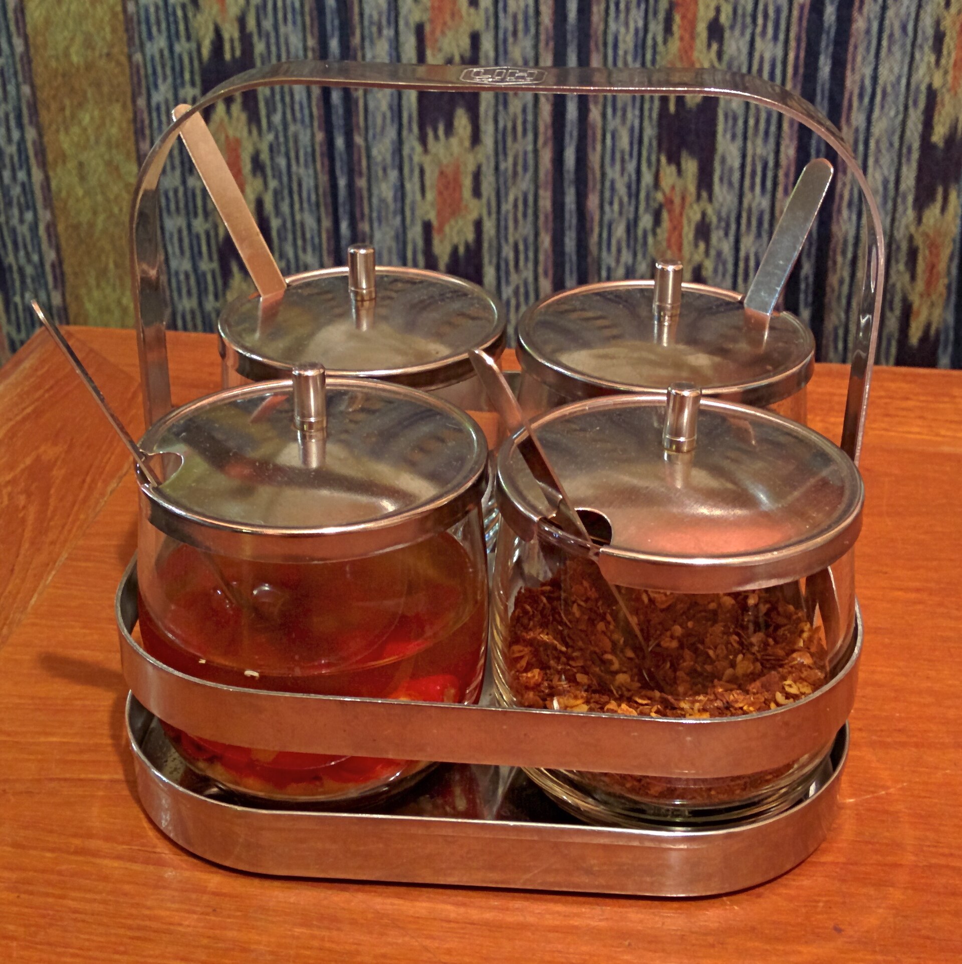 Stainless steel Thai condiment caddy holding four glass pots of chilli in vinegar, dried chilli flakes, sugar and fish sauce with spoons