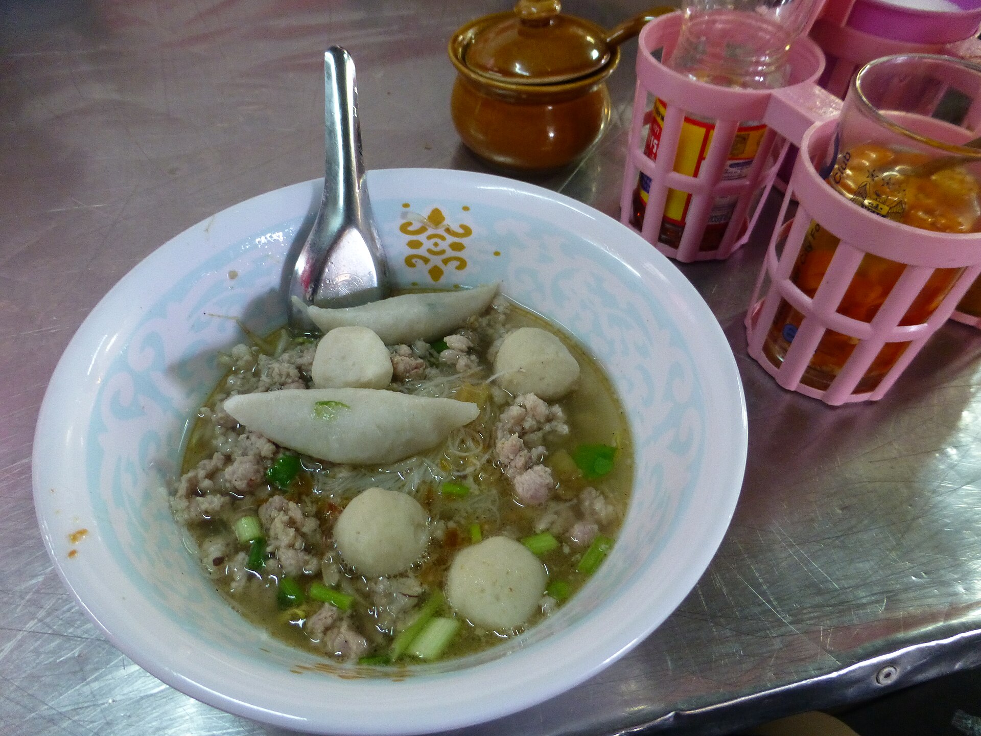 Bowl of Thai kuaitiao nam noodle soup with fish balls, minced pork and spring onion beside a pink condiment caddy on a steel table in Thailand