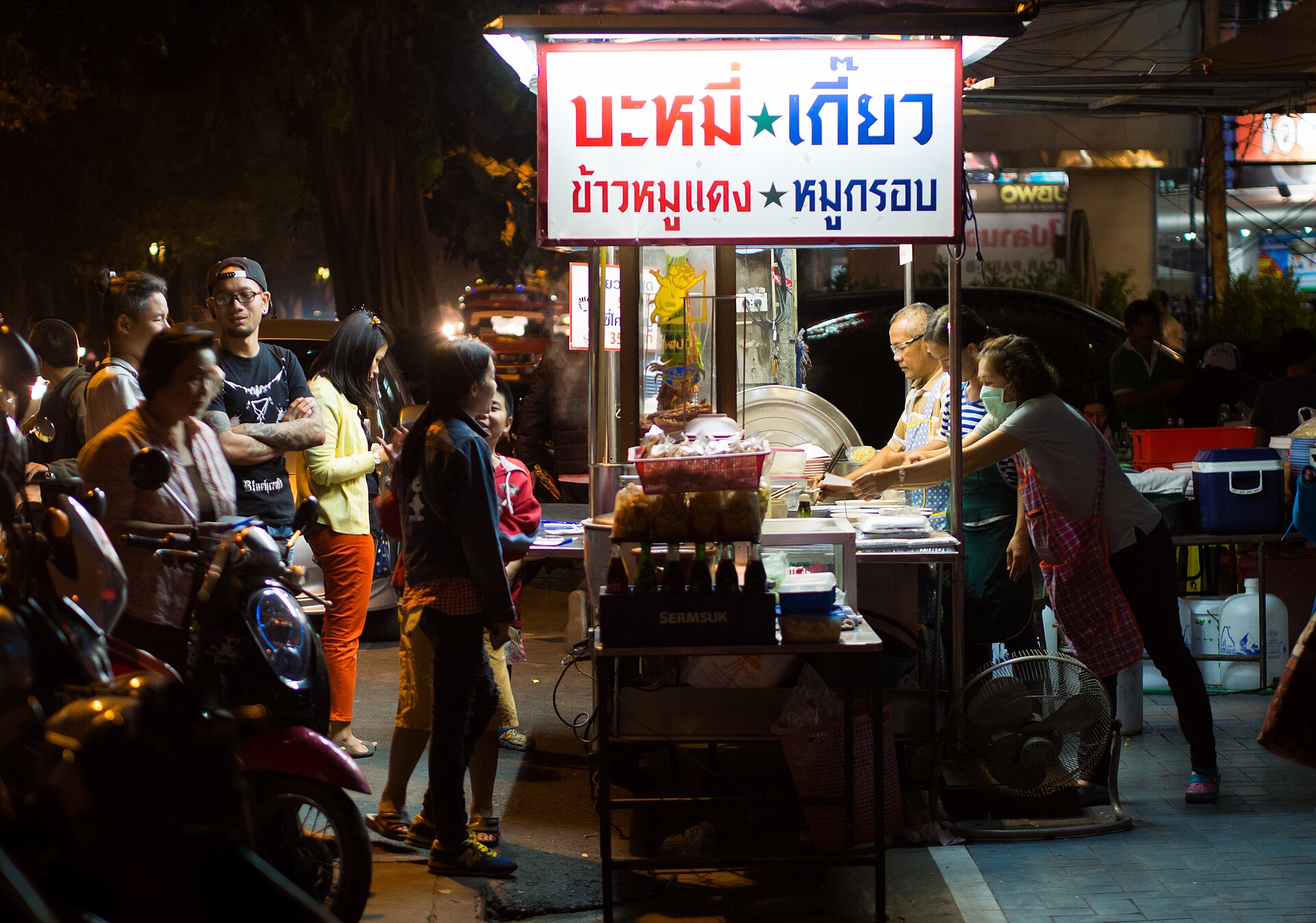 Night-time Thai noodle cart in Chiang Mai with an illuminated red and blue sign reading bamee kiao and khao mu daeng mu krop in Thai script