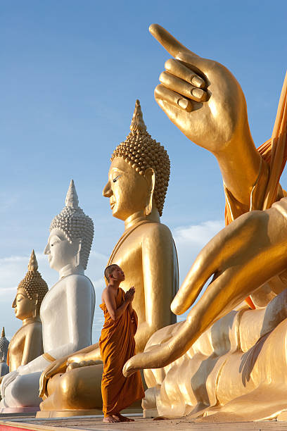 A monk in saffron robes stands with palms pressed together in front of a row of enormous seated Buddha images, gold and white, against a blue sky.